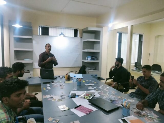 Team members seated around a meeting table while a trainer speaks beside a whiteboard.